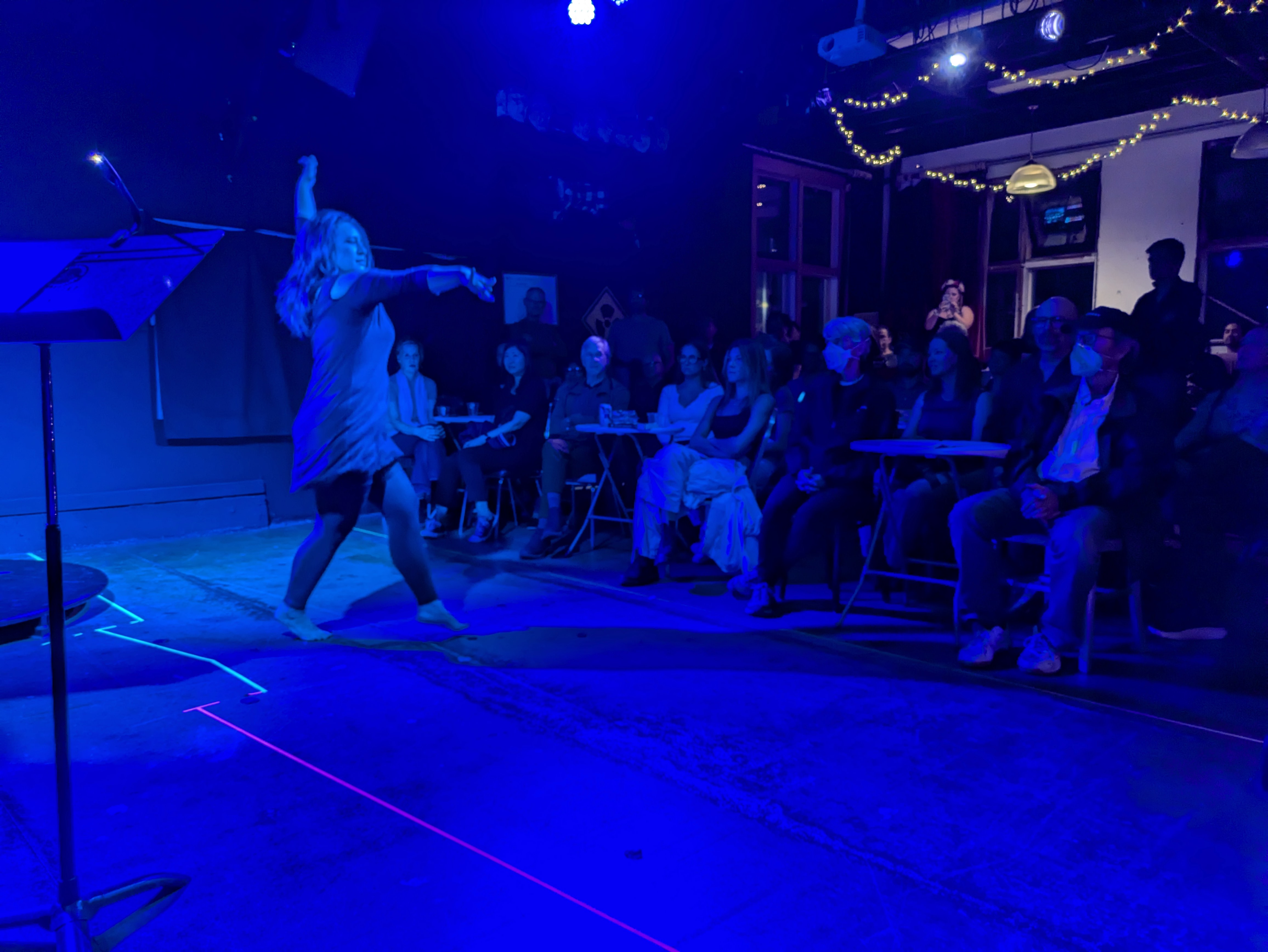 A woman bathed in blue light dances on a makeshift stage before a seated audience.